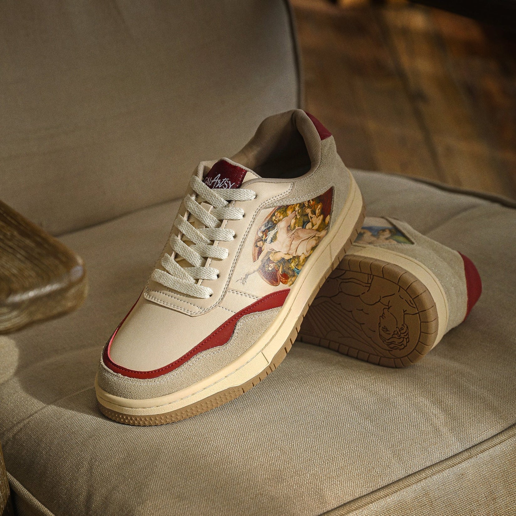 A pair of beige sneakers with red accents and artistic side panels on a couch.